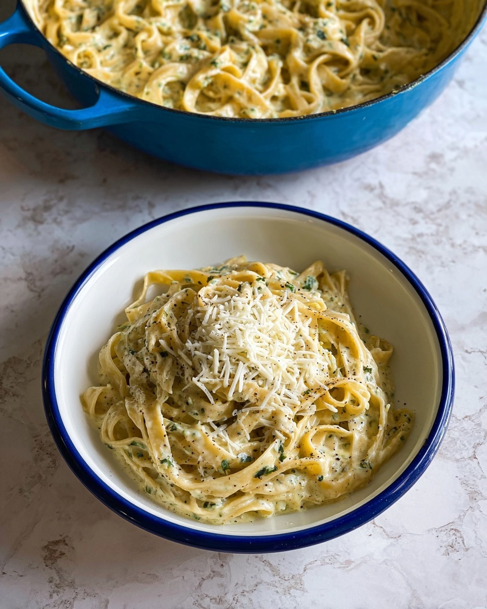 A white bowl filled with creamy fettuccine pasta is placed on a white marbled surface. The pasta has two layers: the bottom layer shows thick, smooth noodles coated lightly in a white cream sauce mixed with small green herb bits, while the top layer is a smaller mound of finely shredded white cheese sitting in the center. The pasta is lightly speckled with black pepper and extra green herbs. Above the bowl, a blue pot filled with more creamy fettuccine pasta is partially visible, the same thick noodles and sauce texture as in the bowl. photo taken with an iphone --ar 4:5 --v 7