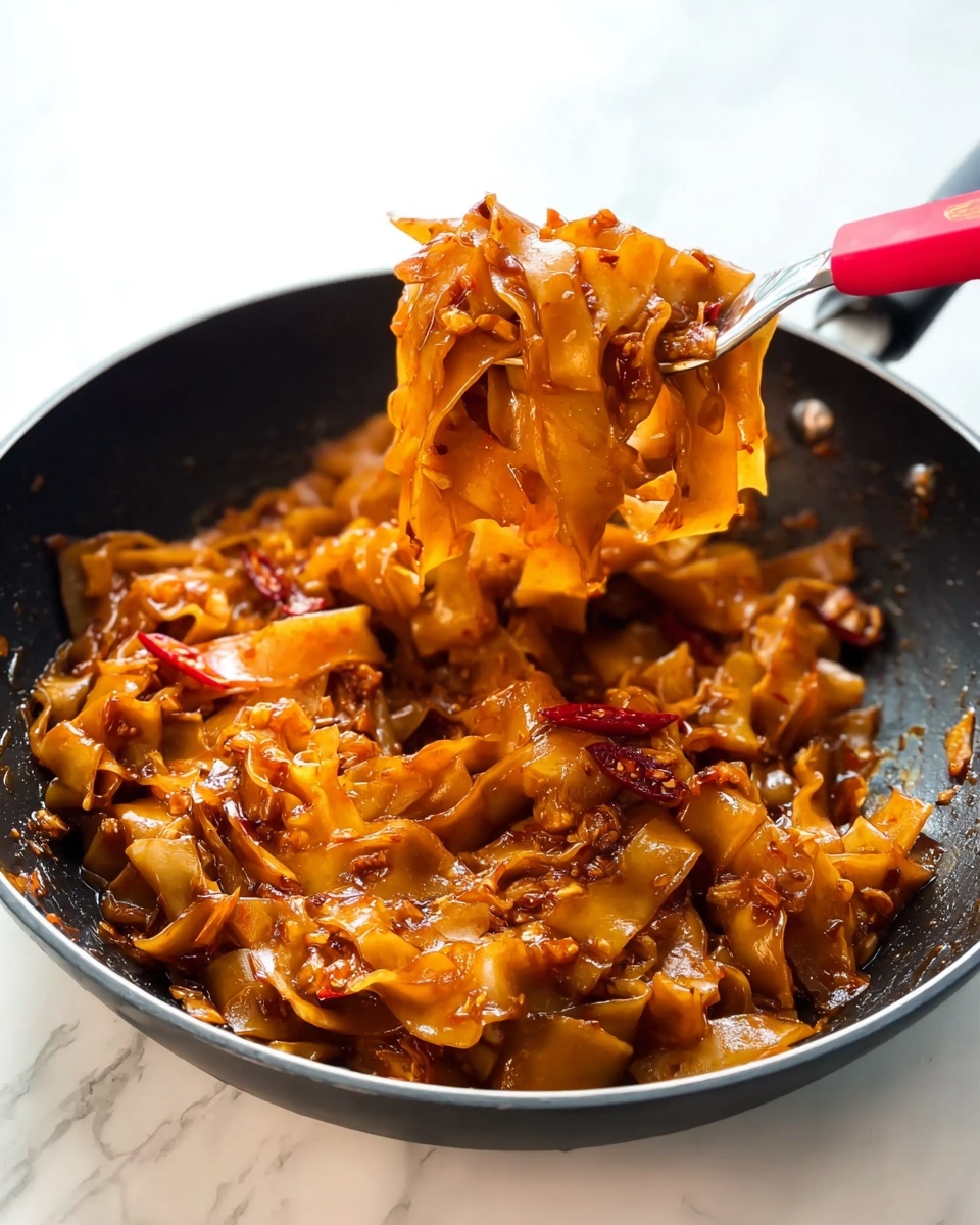 The image shows a black pan filled with wide, flat noodles covered in a thick, rich brown sauce. The noodles are unevenly layered, with some edges curling slightly, and the sauce has a shiny, glossy texture. Scattered on top are thin slices of bright red chili peppers and small white sesame seeds, adding contrast and detail. The pan is placed on a white marbled surface, giving a clean and bright background to the dark pan and colorful food. photo taken with an iphone --ar 4:5 --v 7