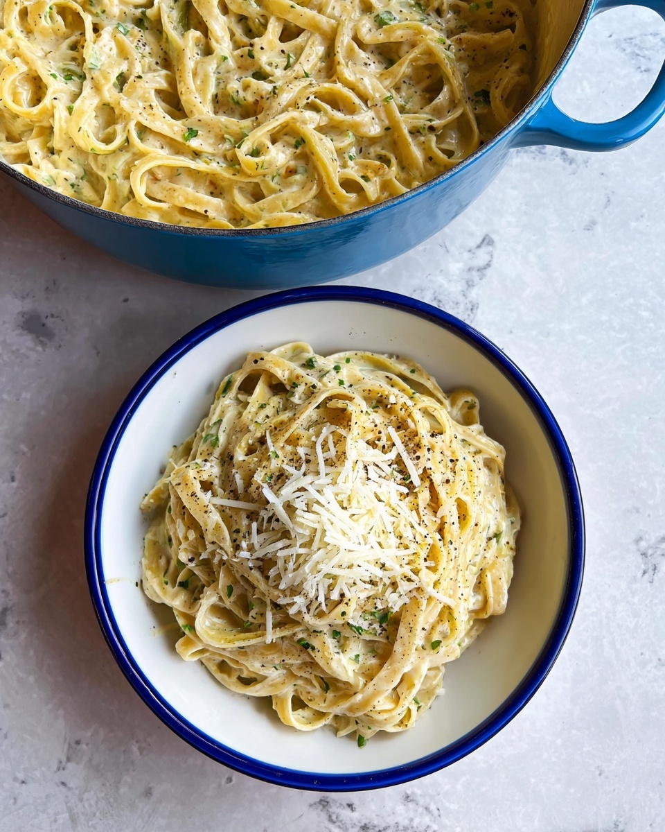 Fettuccine Alfredo Recipe 6 A white plate with light blue edges holds creamy yellow fettuccine pasta coated evenly in a smooth white sauce speckled with green herbs. On top of the pasta is a small mound of finely grated white cheese and scattered black pepper bits. The plate sits on a white marbled surface, and blurred in the background is a blue pot with more pasta inside. photo taken with an iphone --ar 4:5 --v 7