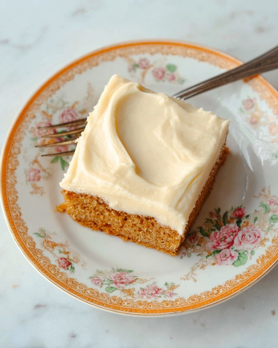 A single square piece of moist brown cake with a light, slightly crumbly texture forms the bottom layer, topped by a thick, smooth layer of creamy white frosting spread evenly across the surface. The cake is placed on a white plate decorated with a delicate floral pattern around the edge. The background is softly blurred with a green apple shape visible in the upper part, and everything sits on a white marbled surface. photo taken with an iphone --ar 4:5 --v 7