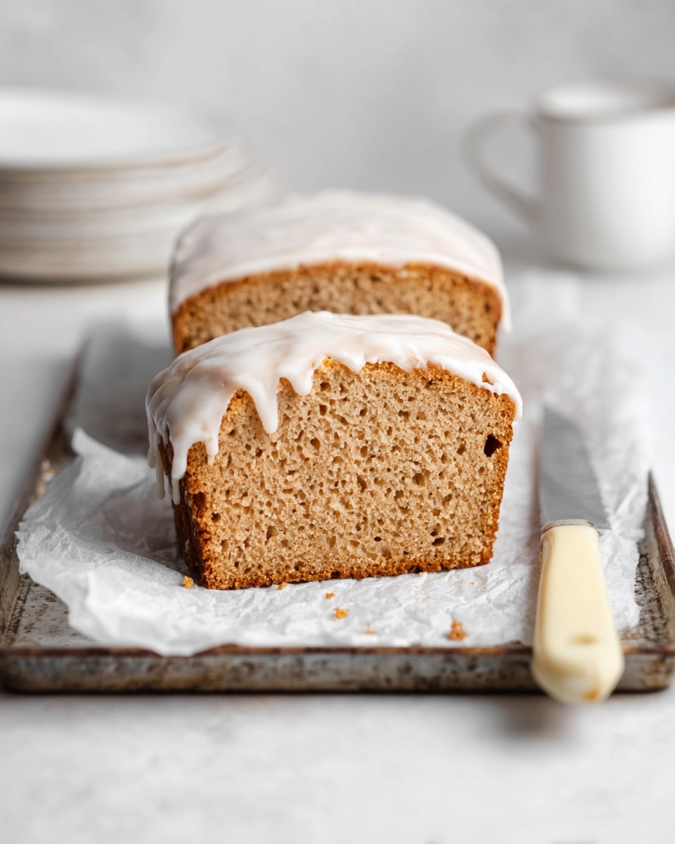 A close-up view of a loaf cake sliced in half, showing two layers of light brown, moist crumb texture with small air holes, topped with a smooth, white glaze that drips down the sides unevenly. The cake rests on a piece of white parchment paper over a rustic gray baking tray, placed on a white marbled surface. To the right of the cake, there is a knife with a beige handle. In the blurred background, there are stacked white plates and a white ceramic cup. Photo taken with an iphone --ar 4:5 --v 7
