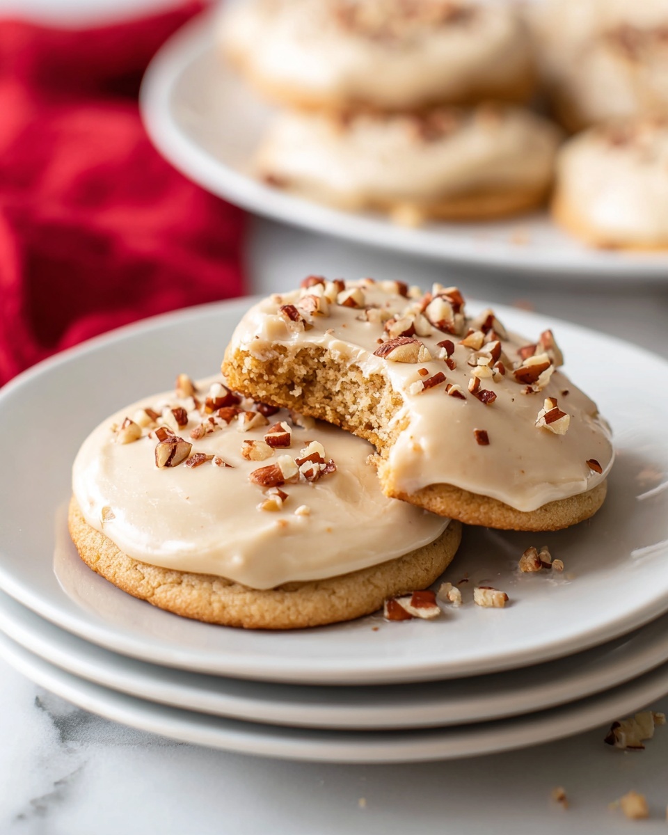 The image shows two round cookies stacked on a white plate, both topped with a thick, smooth layer of light tan frosting. The cookie on the top has a bite taken out of it, revealing a soft, crumbly inside. The frosting is sprinkled with small, chopped pieces of brown nuts, adding texture and color contrast. The plate is placed on a stack of similar white plates, all set on a white marbled surface. In the blurry background, more cookies with the same frosting and nut topping rest on another white plate, while a red cloth peeks out on the side. photo taken with an iphone --ar 4:5 --v 7