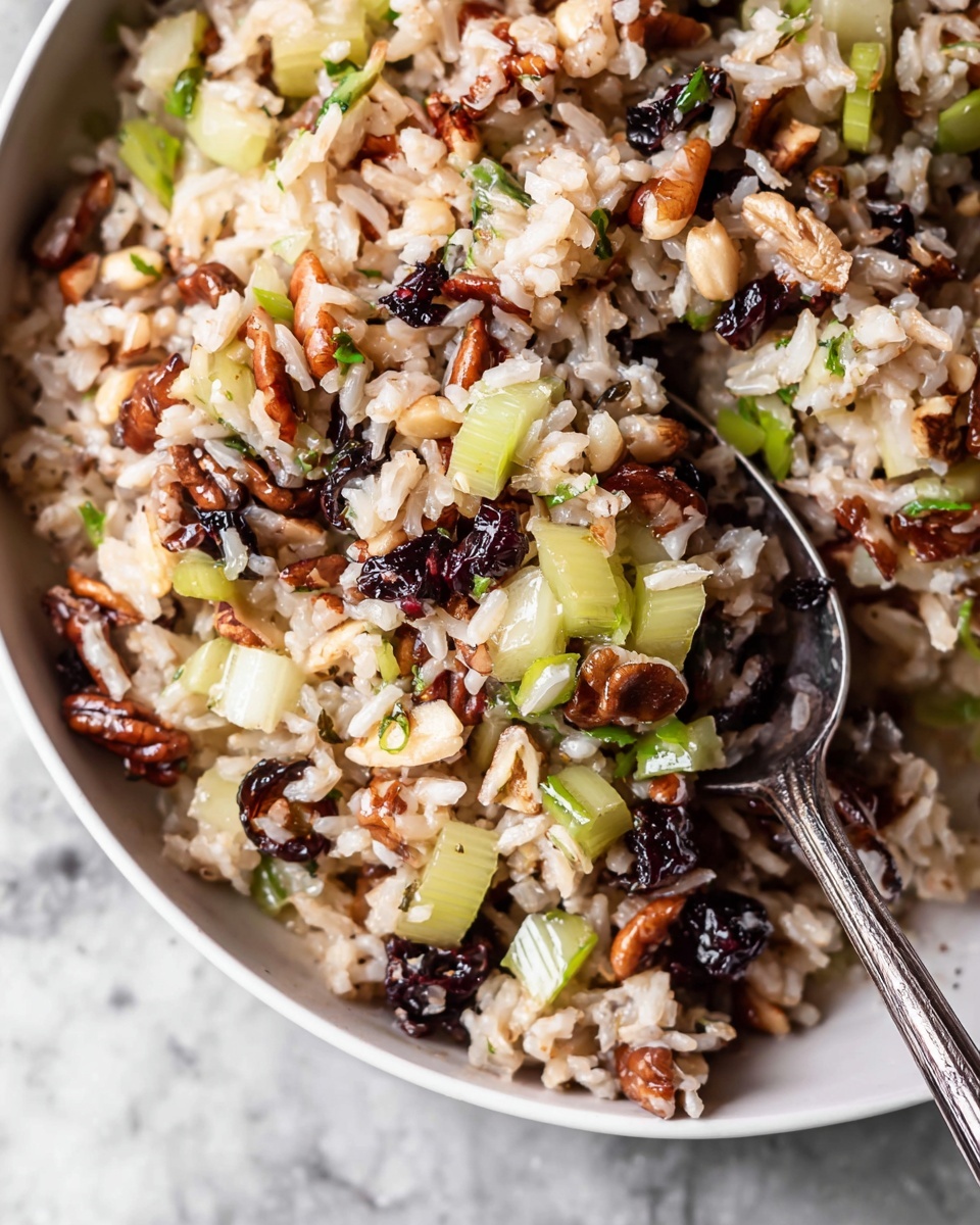 The image shows a close-up of a mixed rice dish in a white bowl with a metal spoon. The dish has many layers and colors including light brown rice, slices of light green celery, dark dried berries, white onion pieces, and brown nuts scattered throughout. The rice looks soft and moist, while the celery and onions add a fresh crunchy texture. The dried berries add dark red and black spots, giving contrast and color to the dish. The metal spoon is partially submerged in the rice mix, ready to serve, all set on a white marbled textured surface. Photo taken with an iphone --ar 4:5 --v 7