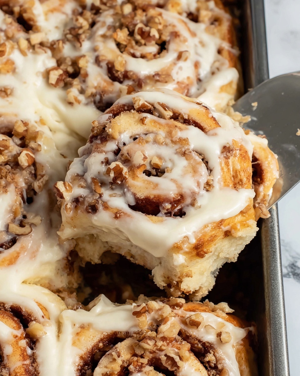 Carrot Cake Cinnamon Rolls Recipe 5 A close-up of a cinnamon roll with three visible layers, each layer showing soft, yellowish dough mixed with bits of orange carrot. Between the layers, there is a dark brown cinnamon-sugar filling that looks sticky and moist. The top layer is covered with a creamy white frosting that spreads about halfway down the sides, sprinkled with small walnut pieces. The roll sits on a white plate with a subtle textured edge, placed on a white marbled surface. Photo taken with an iphone --ar 4:5 --v 7