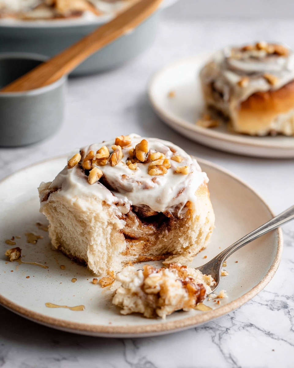The image shows a piece of cinnamon roll on a white plate with three visible layers: the bottom is a soft, light brown dough, the middle has dark brown swirls of cinnamon filling mixed with small pieces of nuts, and the top layer is a thick, creamy white frosting with small walnut pieces scattered on it. The cinnamon roll is partially eaten, revealing the soft texture inside with some cinnamon syrup dripping onto the plate. In the background, there is another white plate with a whole cinnamon roll and a spoon resting beside it, plus a gray bowl with a wooden spatula inside, all set on a white marbled surface. Photo taken with an iphone --ar 4:5 --v 7