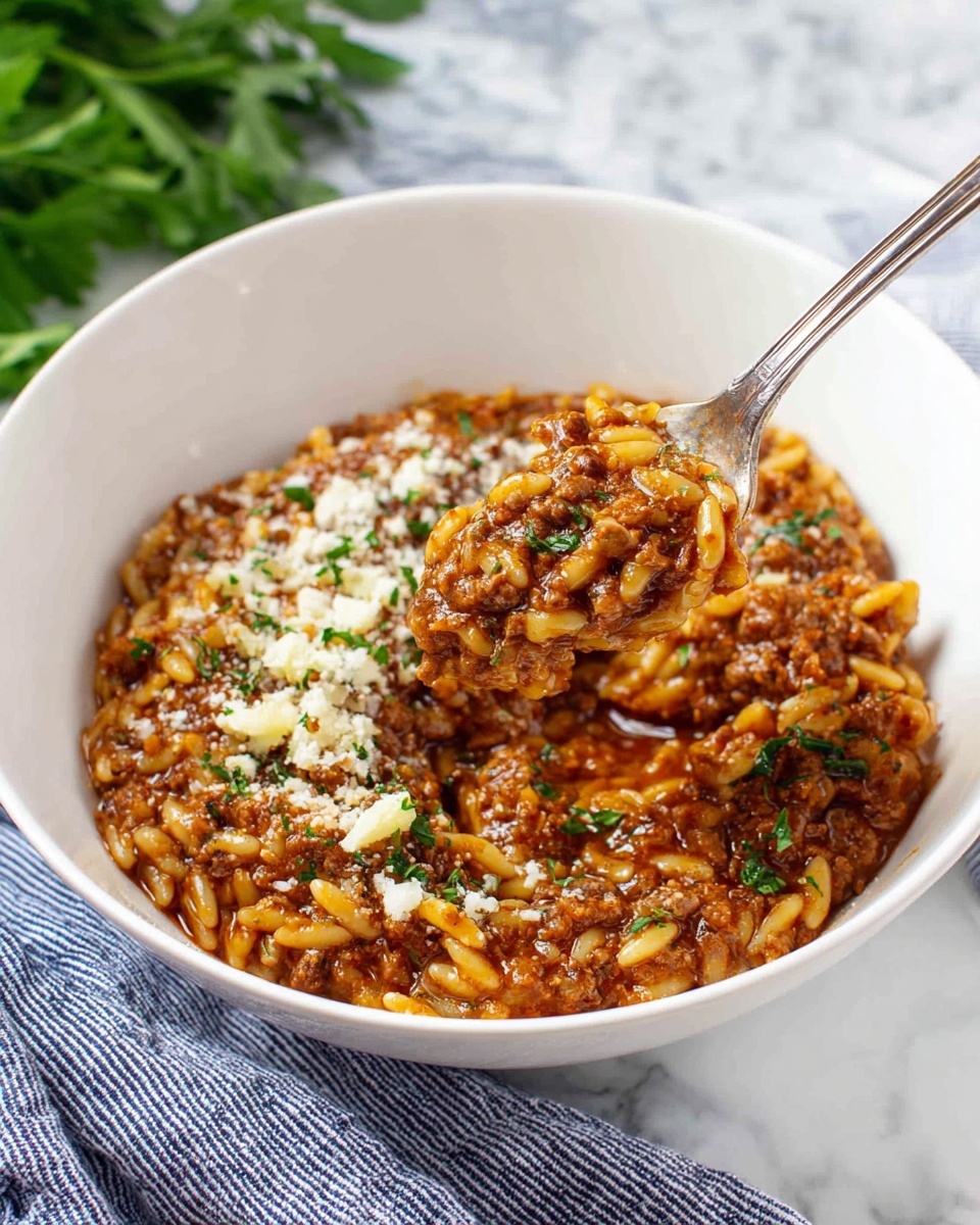 A white bowl holds a rich, thick mix of small pasta in a red-brown sauce filled with ground meat. The pasta mixture is topped with a sprinkle of finely grated cheese and chopped green herbs, which add a fresh contrast. Around the edge of the bowl, there is a thin layer of golden oil or sauce that shines under the light. The bowl sits on a white marbled surface, with a blue and white striped cloth underneath. Fresh green parsley sprigs are scattered around the bowl, adding bright spots of green. Nearby, a silver spoon and fork rest on the cloth, and part of a wooden board with flour and a wooden spoon are visible in the top corner. Photo taken with an iphone --ar 4:5 --v 7