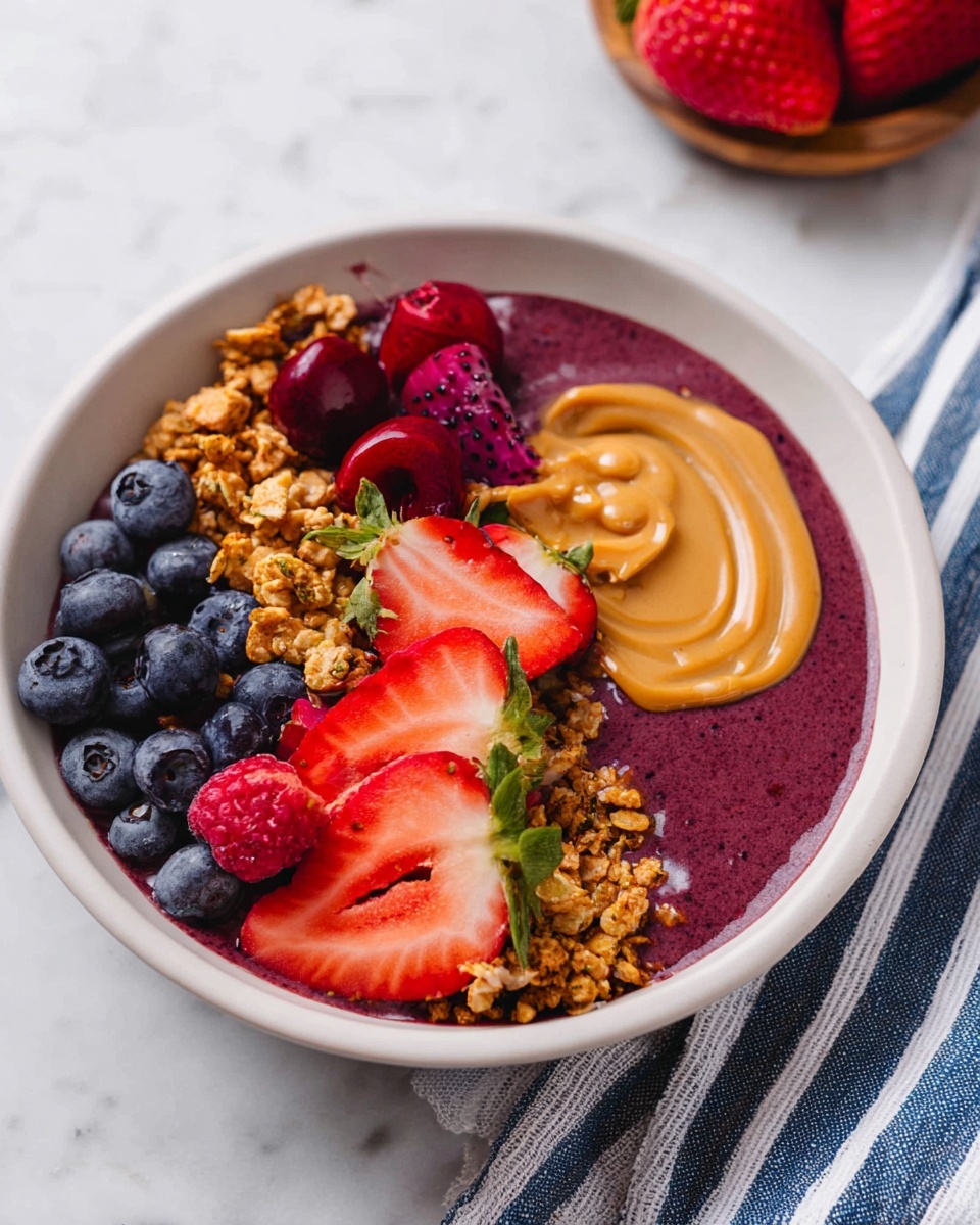A white bowl holds a single serving of a smoothie bowl with a thick, deep purple fruit base visible under toppings arranged on top. The toppings include fresh whole blueberries, sliced red strawberries with green leaves, and halved fresh red cherries. Bright red raspberries and vibrant red dragon fruit pieces with small black seeds add color contrast. Two clusters of golden granola provide a crunchy texture, and a smooth swirl of creamy peanut butter sits in the center. The bowl is placed on a white marbled surface with a striped blue and white cloth nearby. photo taken with an iphone --ar 4:5 --v 7