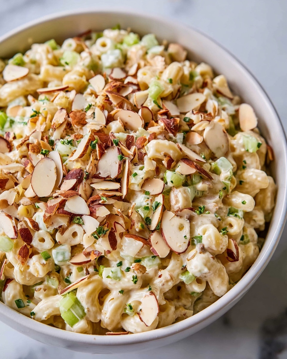A white bowl filled with a creamy macaroni salad that has four visible layers: the base is small elbow macaroni coated in a thick light beige creamy sauce, mixed with small green celery pieces scattered throughout. On top of the macaroni are sliced almonds, browned on the edges with some whole almonds mixed in, adding a crunchy texture. Small bits of green herbs are sprinkled evenly across the dish. The bowl rests on a white marbled surface that adds a clean and fresh feeling to the image. photo taken with an iphone --ar 4:5 --v 7