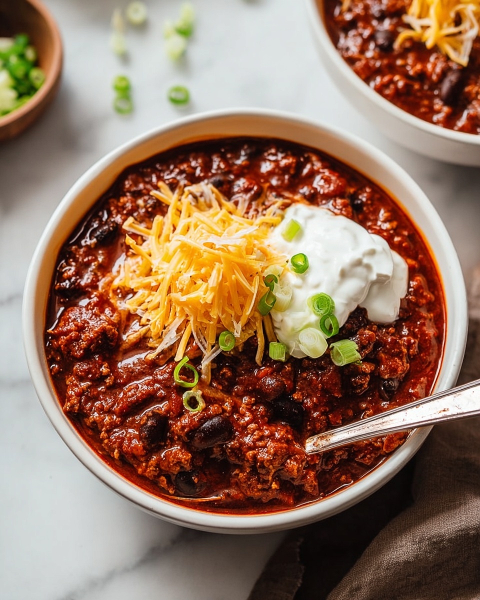 Single white bowl with a hearty serving of rich chili featuring ground beef, black beans, and diced tomatoes in a thick red sauce, topped with a generous mound of shredded cheddar cheese melting slightly, a dollop of smooth sour cream, and fresh sliced green onions for a pop of color, spoon partially immersed revealing the chunky texture, placed on a white marble surface with natural lighting, intimate close-up angled shot emphasizing depth and glossy sauce details, styled like a food blog plated serving photo taken with an iphone --ar 4:5 --v 7