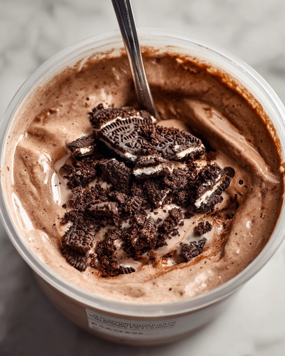 Single white bowl filled with a creamy chocolate milkshake topped with crushed chocolate sandwich cookies, close-up view showing the smooth, thick texture of the shake and the crunchy cookie pieces on top, spoon partially dipped inside, placed on a white marble surface, styled individual serving photo taken with an iphone --ar 4:5 --v 7