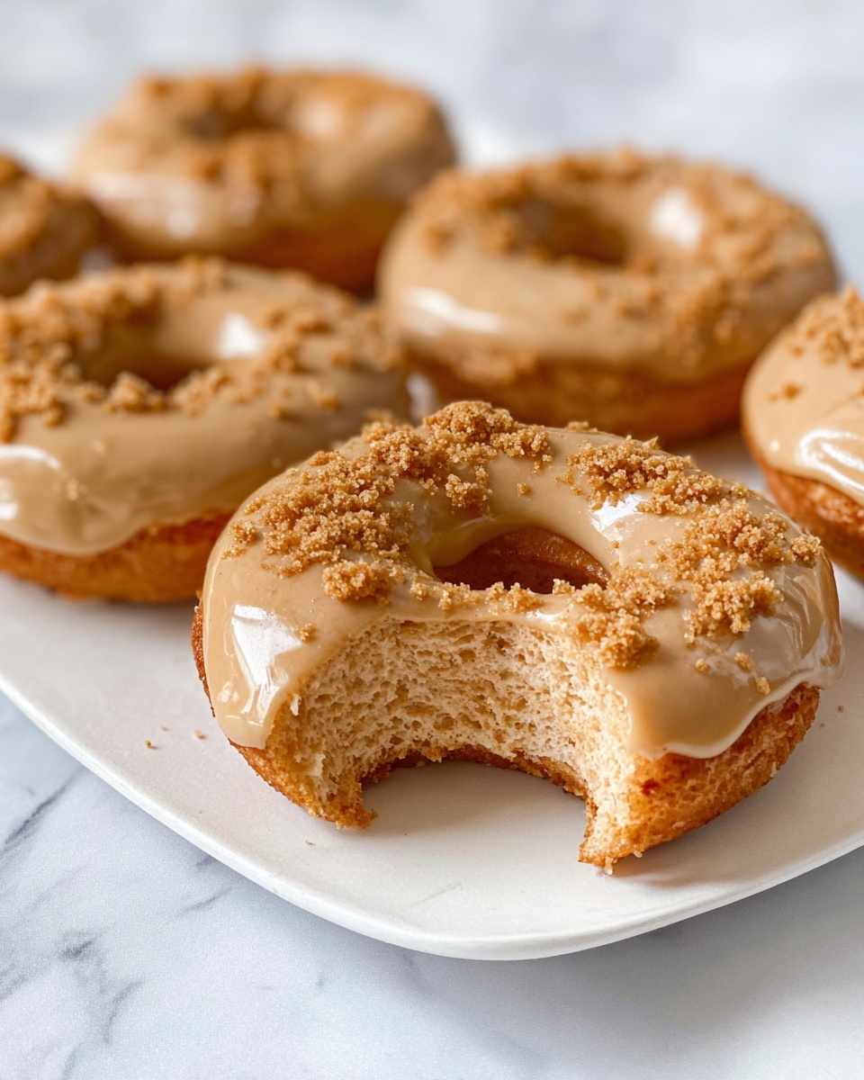 Single white plate with one frosted doughnut featuring a smooth caramel-colored glaze and sprinkled with brown sugar crumbs, showing a visible bite that reveals the soft, airy interior texture, placed on a white marble surface under natural lighting, intimate close-up angled view emphasizing the doughnut’s fluffy crumb and glossy frosting, styled like a food blog serving photo taken with an iphone --ar 4:5 --v 7