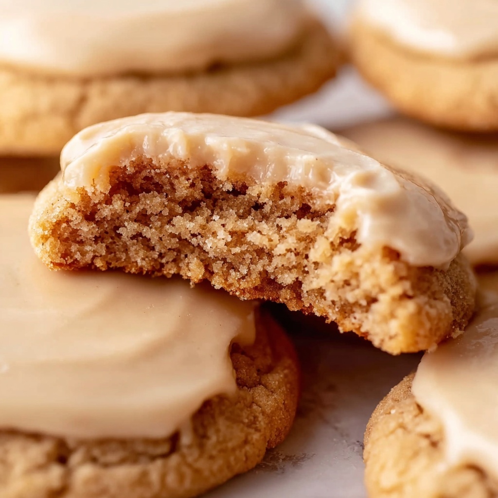 A close-up view of a soft cookie with a light brown color and a crumbly, moist texture shown in layers inside the bite mark. The top of the cookie is covered with a smooth layer of light tan frosting that slightly drips over the edge. The cookie rests among other similar cookies with the same frosting on a white marbled surface, creating a cozy and inviting look. The image is sharp and detailed, showing the texture of both the cookie and frosting clearly. Photo taken with an iphone
