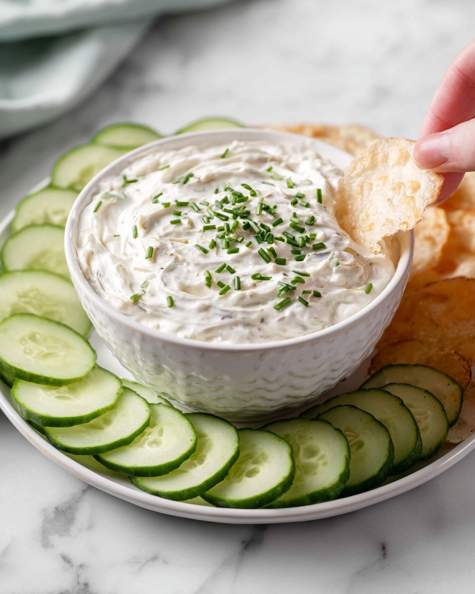 Classic Chip Dip Recipe 6 Large white bowl filled with creamy onion dip garnished with finely chopped fresh chives, surrounded by neatly arranged slices of fresh cucumber on a matching white plate, set on a white marble countertop, shot in natural lighting from a 3/4 angle showcasing the entire bowl and accompaniments, styled like a hero shot from a food magazine, photo taken with an iphone --ar 4:5 --v 7