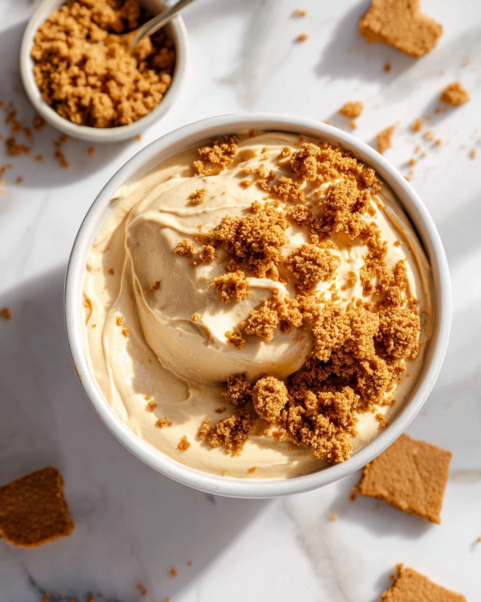 Large white bowl filled with creamy, smooth peanut butter mousse topped generously with crunchy peanut butter cookie crumbles scattered evenly across the surface, accompanied by additional cookie pieces on the side, all set on a pristine white marble countertop, natural light highlighting the dessert's rich texture, professional 3/4 angle food magazine style photo taken with an iphone --ar 4:5 --v 7