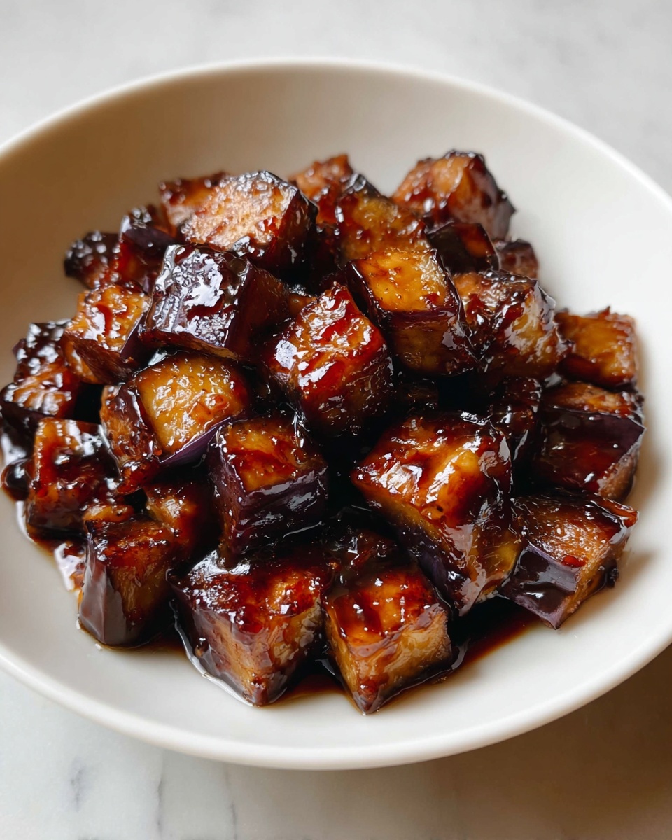 Large white bowl filled with a generous serving of glossy, caramelized cubes of eggplant, each piece perfectly coated in a rich, dark soy glaze that highlights the tender and slightly charred texture of the eggplant skin, arranged to showcase the whole dish under natural lighting on a white marble countertop, professional food magazine hero shot photo taken with an iphone --ar 4:5 --v 7