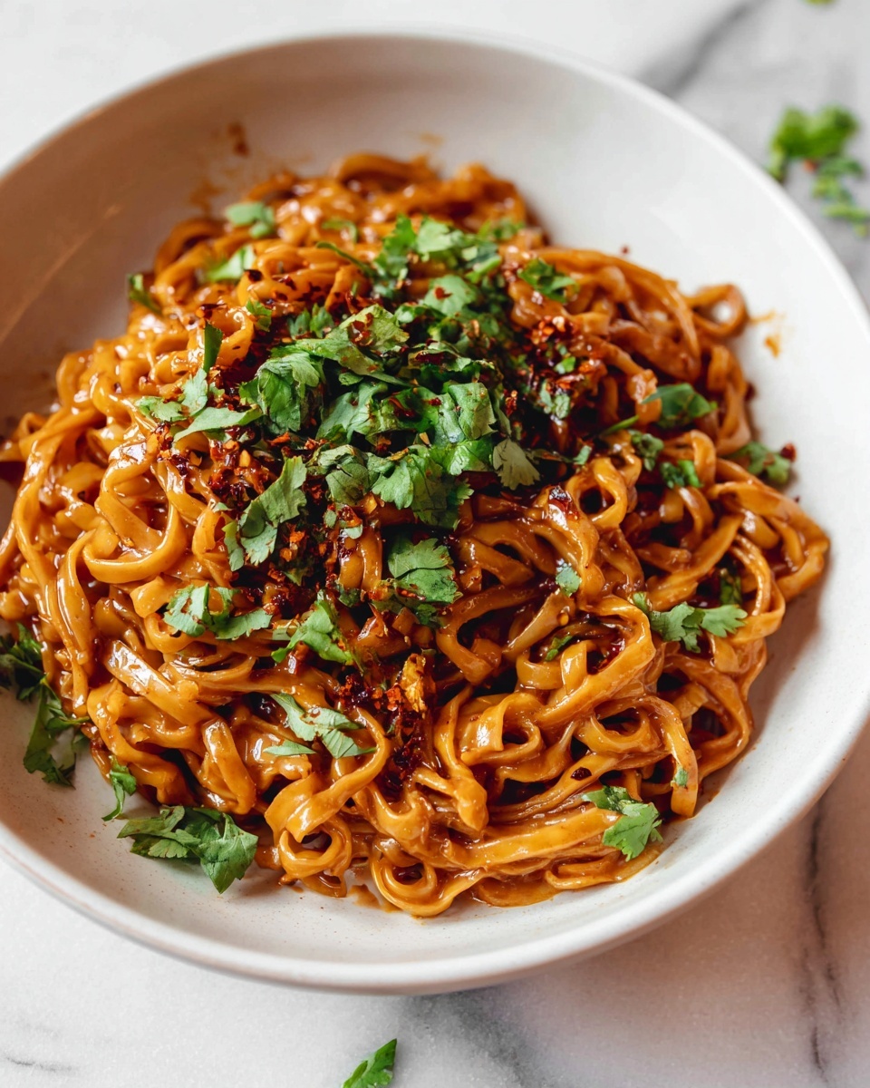 Thai Red Curry Noodles Recipe 6 Large white serving bowl filled with a generous portion of spicy peanut noodles, noodles coated evenly in a rich, creamy orange peanut sauce, garnished with plenty of chopped fresh cilantro and red chili flakes sprinkled on top, the entire dish displayed from a 3/4 angle on a white marble surface under natural lighting, styled as a professional food magazine hero shot, photo taken with an iphone --ar 4:5 --v 7