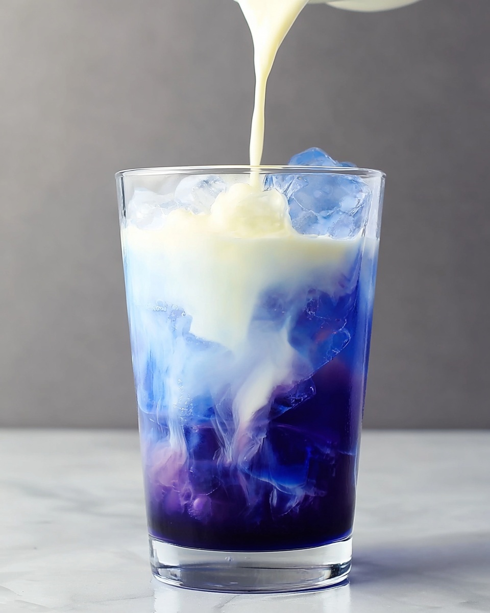 Large white glass filled with vibrant blue butterfly pea flower tea over ice cubes, topped with creamy milk being poured in, showcasing a beautiful swirling gradient from deep blue to milky white, the entire refreshing drink captured in a professional 3/4 angle shot on a white marble background with natural lighting, styled like a food magazine hero shot, photo taken with an iphone --ar 4:5 --v 7